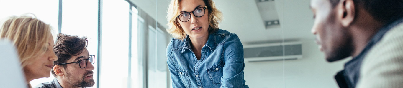 standing woman with glasses in office setting talking to co-workers that are sitting